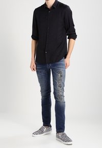 Uomo che indossa una camicia nera a maniche lunghe con bottoni, jeans blu strappati con toppe e scarpe slip-on con stampa zebrate bianco e nere, in piedi su uno sfondo bianco.