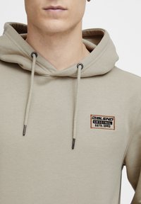 Beige hoodie met trekkoorden, met een rechthoekig geborduurd logo in zwart en oranje. Zachte textuur en casual ontwerp.