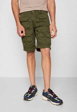 Uomo che indossa pantaloncini cargo verde oliva con multiple tasche a pattina e sneakers atletiche multicolori, in piedi su un pavimento grigio.