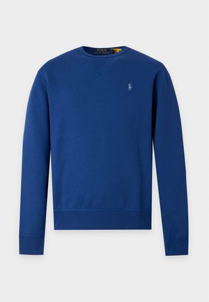 Homme portant un sweat-shirt bleu assorti et un pantalon de survêtement avec des baskets blanches, debout devant un fond clair uni.