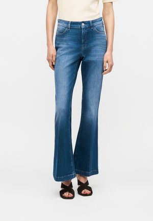 Frau trägt blaue Jeans mit mittlerer Leibhöhe und Schlag sowie schwarze Sandalen mit gekreuzten Riemen, stehend vor einem schlichten weißen Hintergrund.