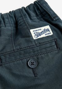 Donkerdenim broek met elastische tailleband, voorzien van een wit label met de tekst "Superdry" en een zwarte knop met een logo op de voorkant van de zak.