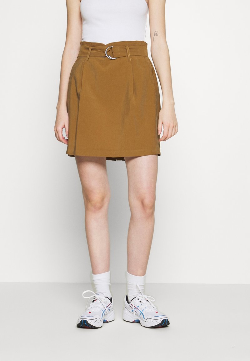 Falda de lona marrón con un cinturón que presenta una hebilla circular de plata, dos bolsillos laterales y un diseño de pliegues, combinada con calcetines blancos y zapatillas deportivas.