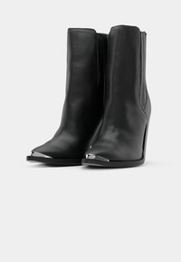 Botines de cuero negro hasta el tobillo con punta afilada, acento plateado brillante en la punta y un alto tacón de aguja. Textura suave y diseño elegante.