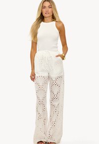 Witte mouwloze top met een hoge halslijn en uitgesneden armopeningen, gecombineerd met losse witte broeken met ingewikkelde oogjespatronen en een trekkoord in de taille.