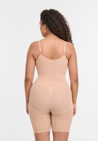 Kvinna med lockigt hår iförda beige shapewear-shorts och linne, sedd bakifrån mot en enkel vit bakgrund.