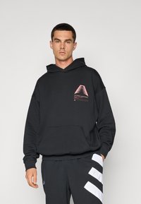 Fekete, pamutkeverékből készült kapucnis pulóver, amelyen rózsaszín geometrikus logó díszíti a mellrészt, kenguruzsebbel és bordázott mandzsettákkal. Sötét nadrággal párosítva.