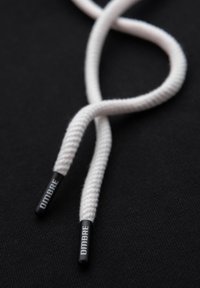 Bílé tkaničky s copánkovou texturou, se černými plastovými konci s nápisem "OMBRE", na černém pozadí.