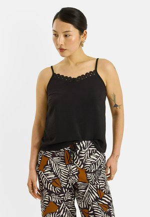 Femme aux cheveux foncés portant un caraco noir orné de dentelle et un pantalon à motifs de feuilles marron et blanches, debout devant un fond uni.