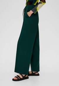 Green wide-leg pants with a drawstring waistband, side pockets, and smooth texture, paired with black sandals featuring a strappy design.