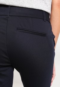 Navy kalhoty s úzkým střihem a hladkou texturou, které mají jednu zadní kapsu a jemné prošívání. Materiál má mírný lesk.