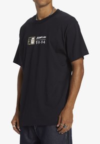 Black cotton t-shirt featuring a graphic logo in green and white, crew neckline, relaxed fit, and short sleeves.