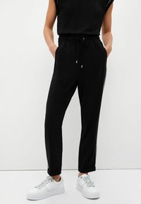 Czarne spodnie dresowe z bocznymi zamkami, elastycznym pasem i sznurkiem. Połączone z białymi sneakersami z teksturowanymi sznurowadłami.