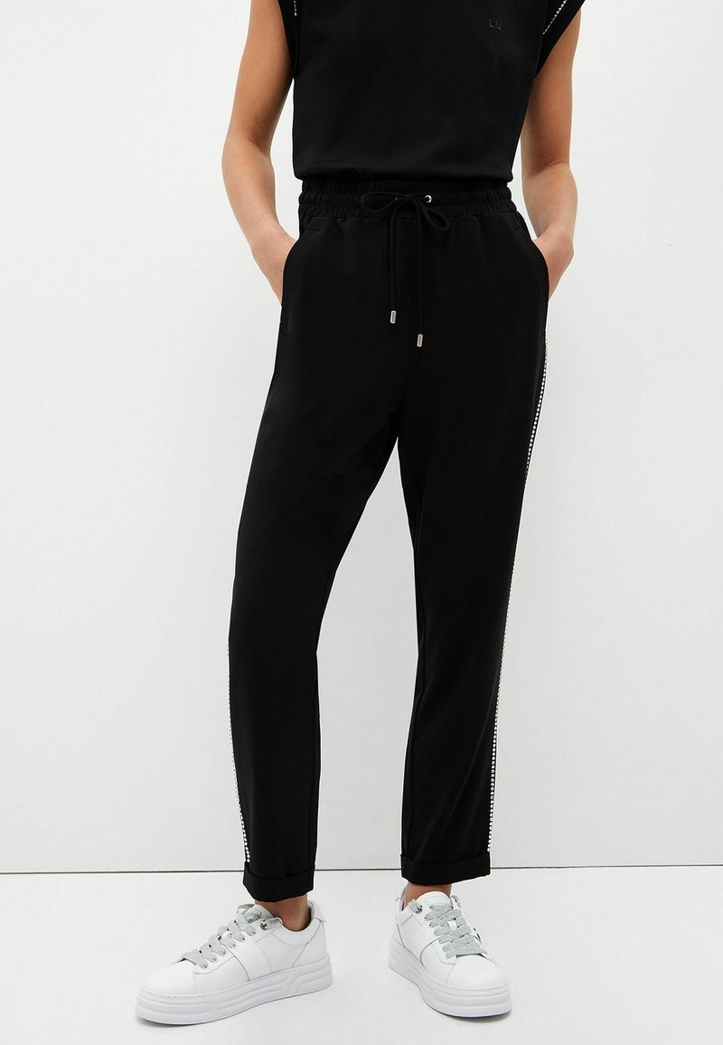 Czarne spodnie dresowe z bocznymi zamkami, elastycznym pasem i sznurkiem. Połączone z białymi sneakersami z teksturowanymi sznurowadłami.