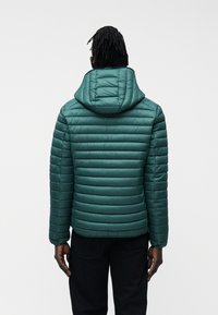 Άνδρας με σκούρο δέρμα και κοτσιδάκια, φοράει μια σακάκι με κουκούλα σε τιρκουάζ χρώμα με ραμμένα σχέδια και μαύρο παντελόνι, όπως φαίνεται από πίσω μπροστά σε λευκό φόντο.