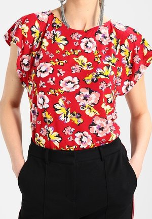 Frau trägt eine rote kurzärmelige Bluse mit Blumenmuster, die in eine hoch taillierte schwarze Hose gesteckt ist, mit Händen in den Taschen.