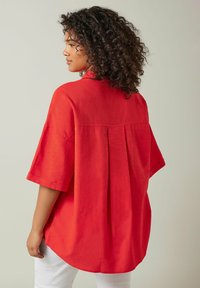 Camisa roja oversize hecha de tejido ligero, con mangas cortas, cuello de solapa y un diseño con pliegues en la parte posterior.