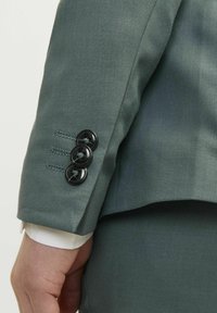 Manche de veste de costume vert foncé avec trois boutons noirs et un revers blanc contrasté. La texture du tissu semble lisse et ajustée.