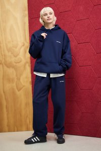 Completo felpa e pantaloni della tuta blu navy, con cappuccio, coulisse e dettaglio del logo bianco. Il modello indossa scarpe nere con strisce bianche.