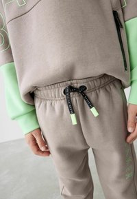 Helle graue Sweatshirt mit grünen Akzenten und Tasche, kombiniert mit passenden hellgrauen Sweatpants mit Kordelzug und "BAKER"-Logo.