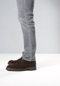 Braune Wildleder-Stiefeletten mit strukturierter Oberfläche und Schnürdesign, kombiniert mit eng anliegenden grauen Jeans, Seitenansicht.