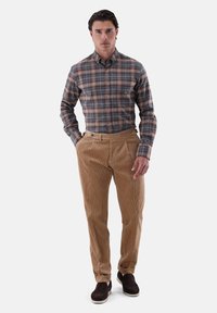 Karierte Hemd in Grau-, Braun- und Blautönen; hellbraune Cordhosen mit strukturierter Oberfläche; braune Slipper; minimaler Schmuck.