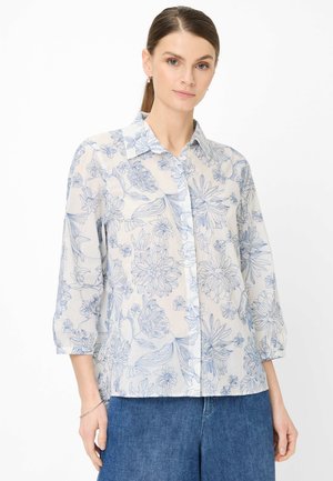 Frau trägt ein weißes Hemd mit blauen Blumenmuster und dreiviertellangen Ärmeln, kombiniert mit blauen Jeans, steht ruhig da.