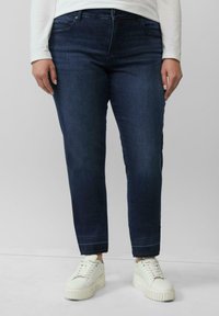 Dunkelblaue Denim-Jeans mit geradem Bein, ausgestattet mit Fronttaschen und einem rohen Saum. Getragen mit weißen klobigen Turnschuhen.