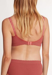 Soutien-gorge en tissu brique côtelé avec bretelles réglables, fermeture à crochets et œillets dans le dos, et bords lisses. Les cheveux du modèle tombent en cascade dans le dos.