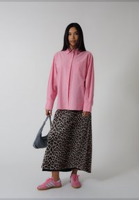 Rosa randig skjorta med knappar, lång leopardmönstrad kjol och rosa sneakers. Håller en grå väska. En enkel bakgrund framhäver outfiten.