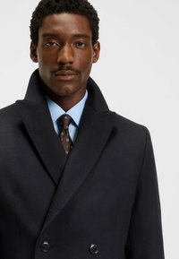 Manteau en laine bleu marine à double boutonnage avec larges revers et deux boutons noirs, associé à une chemise bleu clair et une cravate brune à motifs.
