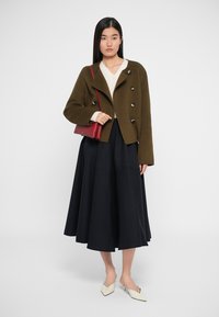 Olive rohelise villase segu jope topelt nööpidega, kreemjas kudum, must plisseeritud midi seelik ja valged terava ninaga kingad.