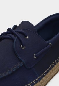 Ναυτικό μπλε σουέτ παπούτσι σκαφάκι με στρογγυλά κορδόνια, μεταλλικά περβάζια και λεπτομέρειες ραφής στη σόλα από πλεγμένο τζούτ.