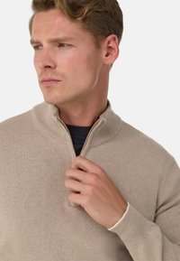 Pull beige à fermeture quart de zip en tissu tricoté doux, avec col et poignets côtelés. Le mannequin tient la fermeture éclair légèrement ouverte.
