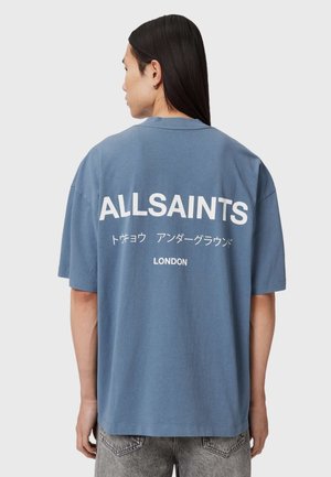 Person mit langen dunklen Haaren trägt ein übergroßes blaues T-Shirt mit weißem "ALLSAINTS", japanischem Text und "LONDON" auf dem Rücken.