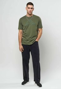 Mann iført olivengrønn t-skjorte med trykk, svarte jeans, svarte joggesko og sølvkjede, stående mot en ensfarget lys bakgrunn.