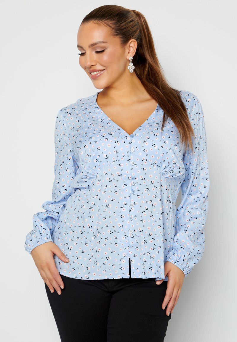 Blusa floreale azzurro chiaro con chiusura a bottoni, maniche lunghe e texture arricciata. Presenta piccoli fiori sparsi in rosa e blu navy.
