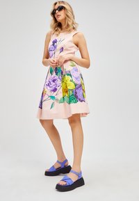 Abito floreale rosa con stampe violetto e giallo, forma a trapezio, design senza maniche. Abbinato a sandali con plateau blu.