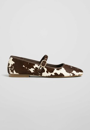 Zapato tipo Mary Jane plano con estampado de vaca marrón y blanco, con una sola correa y hebilla dorada, tacón bajo y punta redondeada.