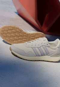 Bílá teniska Adidas s béžovou podrážkou a třemi světle šedými pruhy, jedna bota stojí vzpřímeně a druhá leží na boku, zobrazující vzor podešve.