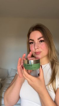 Femme aux cheveux longs tenant un pot vert d'Elemis Pro-Collagen Green Fig Cleansing Balm dans une chambre aux murs neutres.