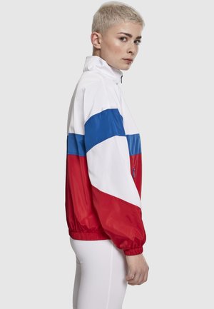 Rövid szőke hajú személy, aki vörös, fehér és kék színblokkokból álló, magas galléros dzsekit visel, oldalról látható.