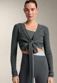 Top corto a maniche lunghe a costine verde scuro con dettaglio annodato sul davanti. Indossato sopra un reggiseno sportivo grigio e blu, abbinato a leggings neri a vita alta.