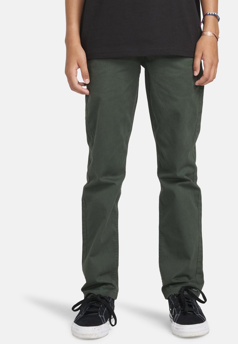 Persoon in donkergroene rechte broek, zwarte sneakers met witte zolen en een zwart shirt, staand op een witte achtergrond.