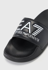 Črne slide sandale z strukturiranim gumijastim podplatom in širokim trakom, na katerem je odtisnjeno belo oblikovanje, vključno z "EA7" in "Emporio Armani."