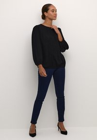 Blouse noire texturée avec des manches bouffantes, associée à un jean skinny bleu foncé et des talons hauts noirs, portée par un mannequin sur fond neutre.