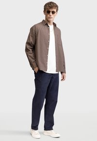 Camisa de cuadros marrón sobre una camiseta blanca, combinada con pantalones azul oscuro y zapatillas beige, con gafas de sol redondas y una cadena como collar.