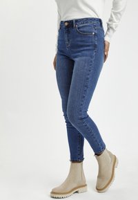 Jean skinny en denim bleu moyen, avec une taille haute et du matériel en cuivre, associés à des bottines beige à semelle épaisse.