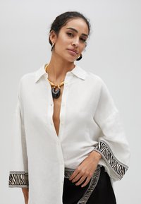 Camicia di lino bianca con maniche larghe, rifinita con un bordo a motivi zebrate. Indossata con una collana gialla e nera e dettagli neri testurizzati.