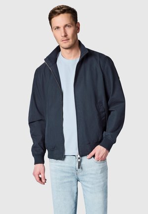 Mand iført en navy lynlåsjakke over en lyseblå skjorte og lyse jeans mod en ensfarvet lys grå baggrund.